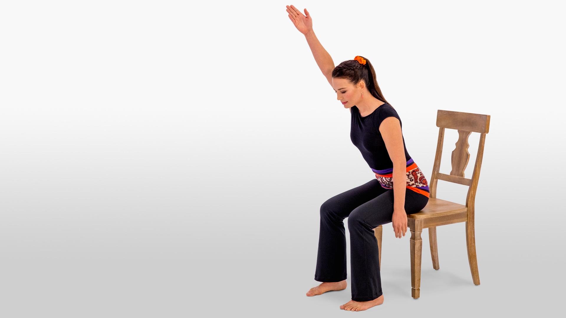 Woman demonstrating a chair yoga pose with arm raised while seated on a sturdy wooden chair